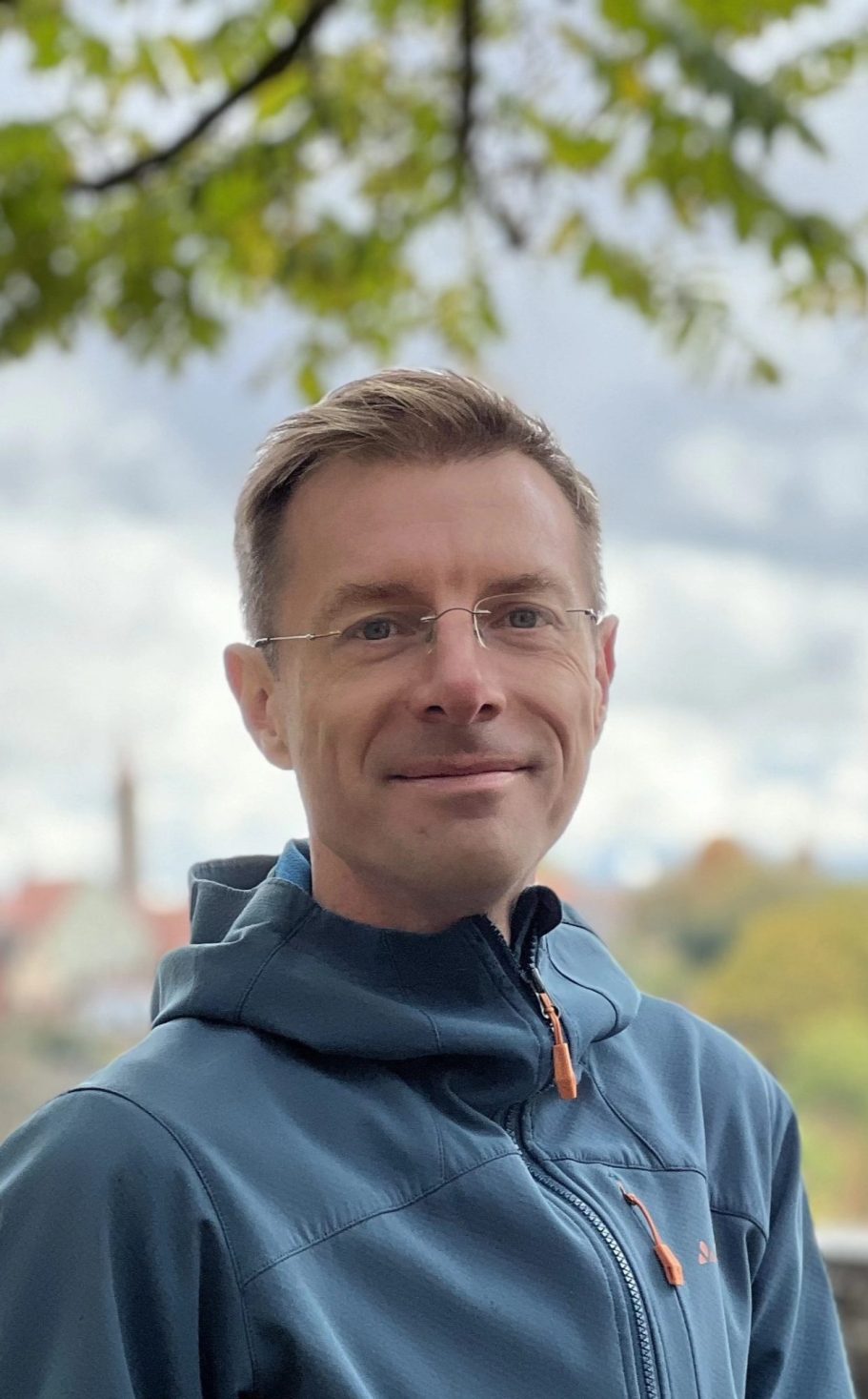 Simon Jarr Mann mit kurzem Haar, lächelnd, trägt eine blaue Jacke, Hintergrund mit Bäumen und Himmel.
